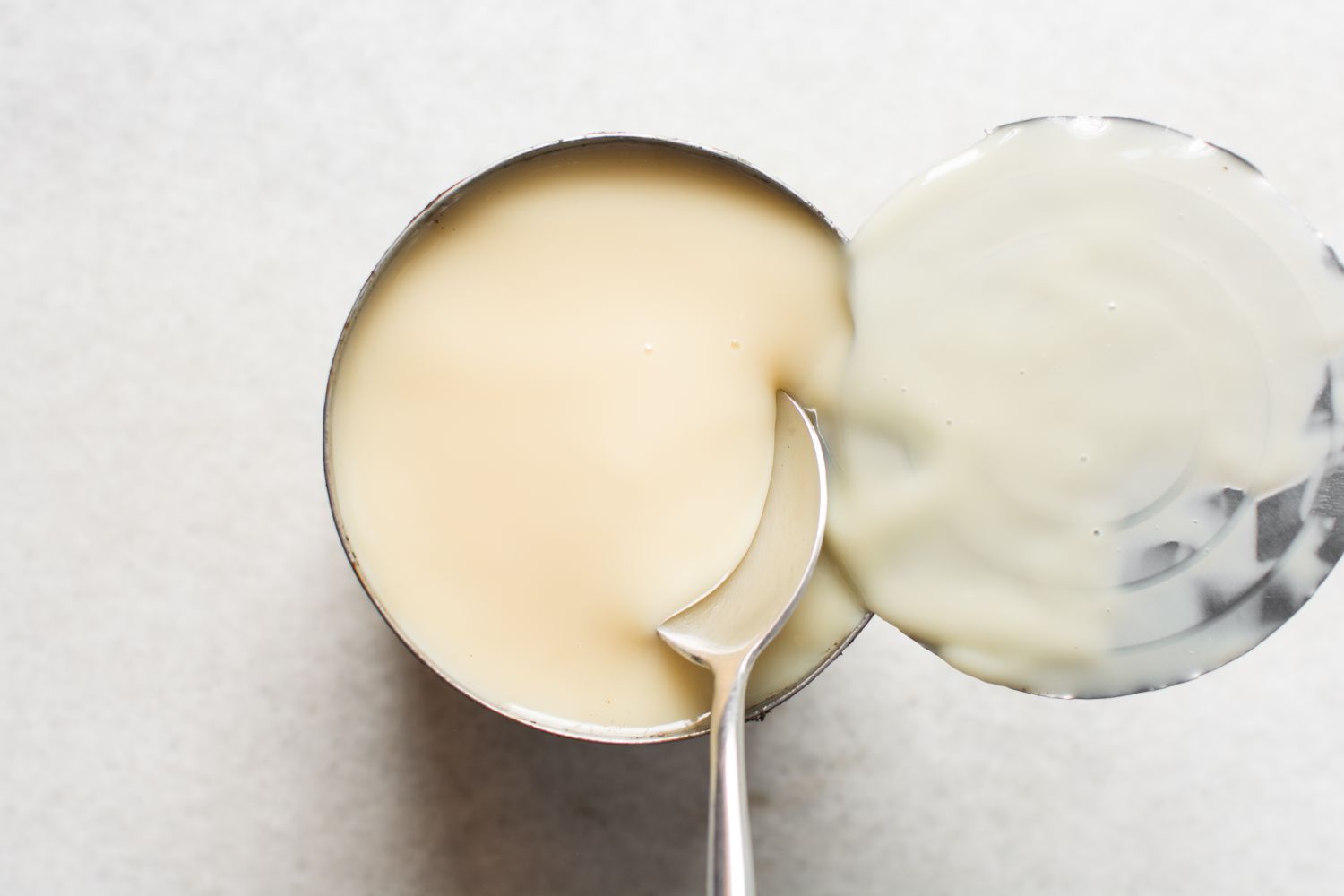 A can of condensed milk with a spoon inside it and the lid partially peeled back positioned on a white surface