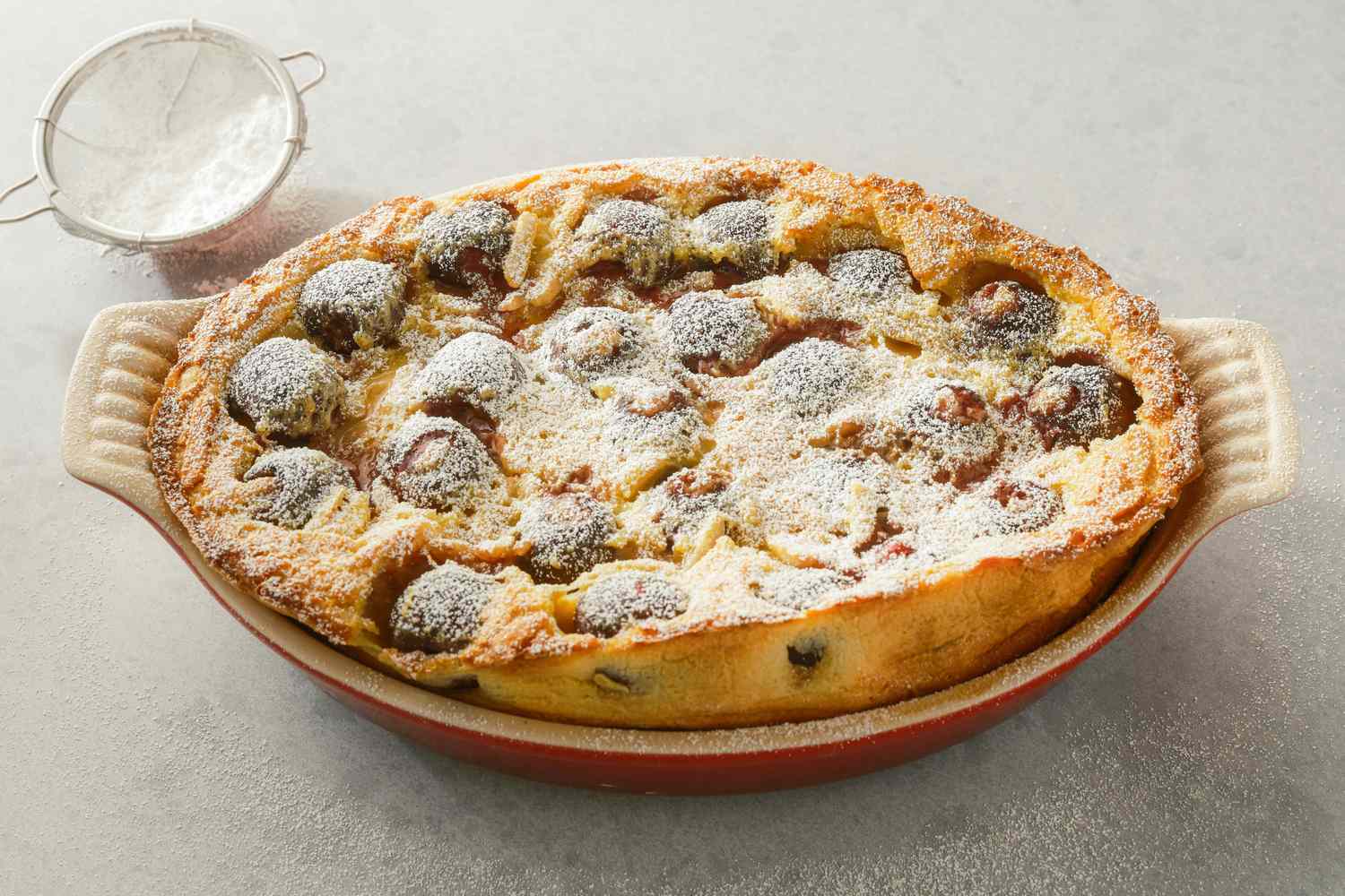 A cherry clafoutis in a baking dish with powdered sugar on top, resting on a surface with a sifter nearby