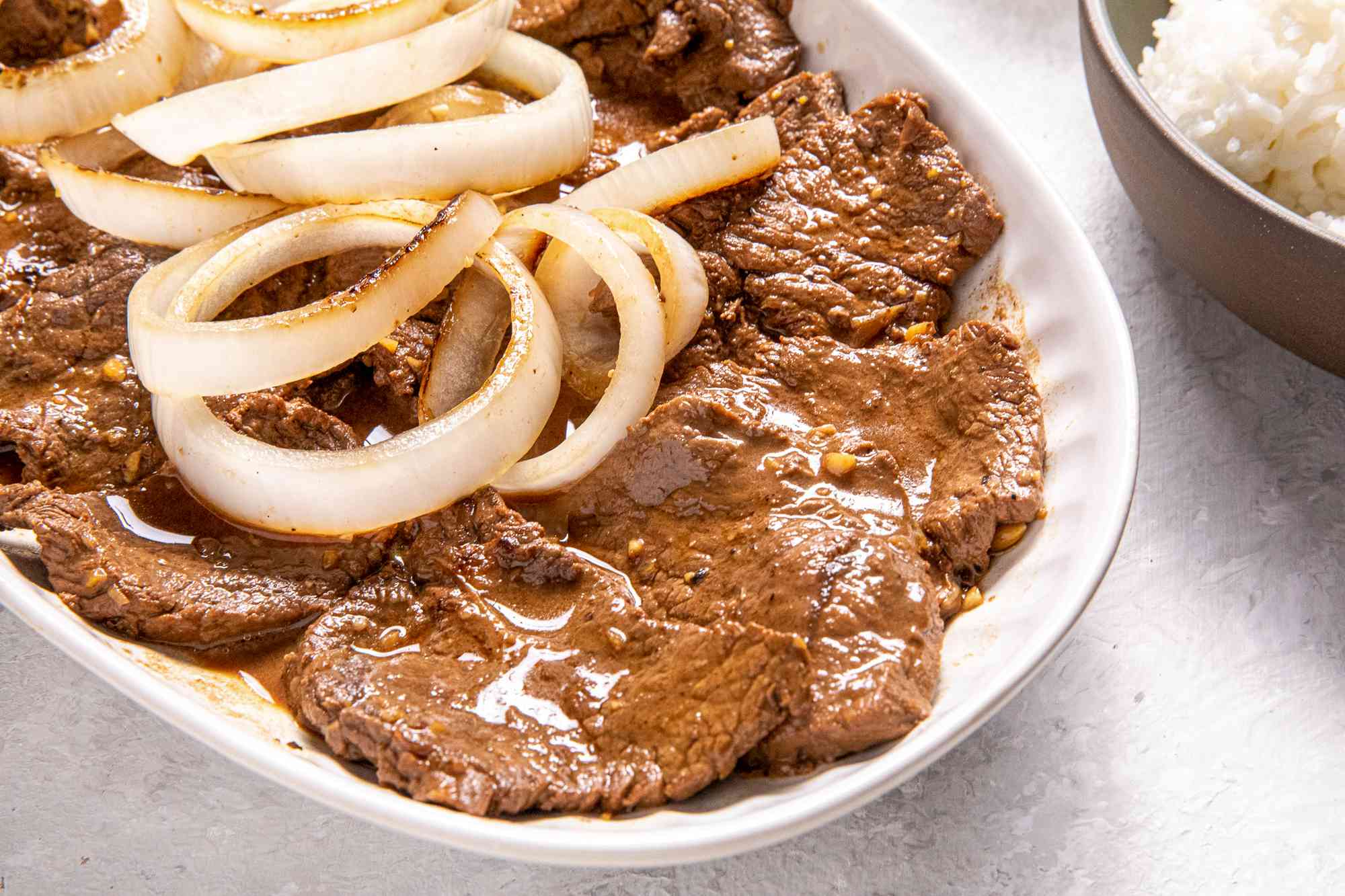 Plate of Bistek Topped With Onions Next to a Bowl of Rice
