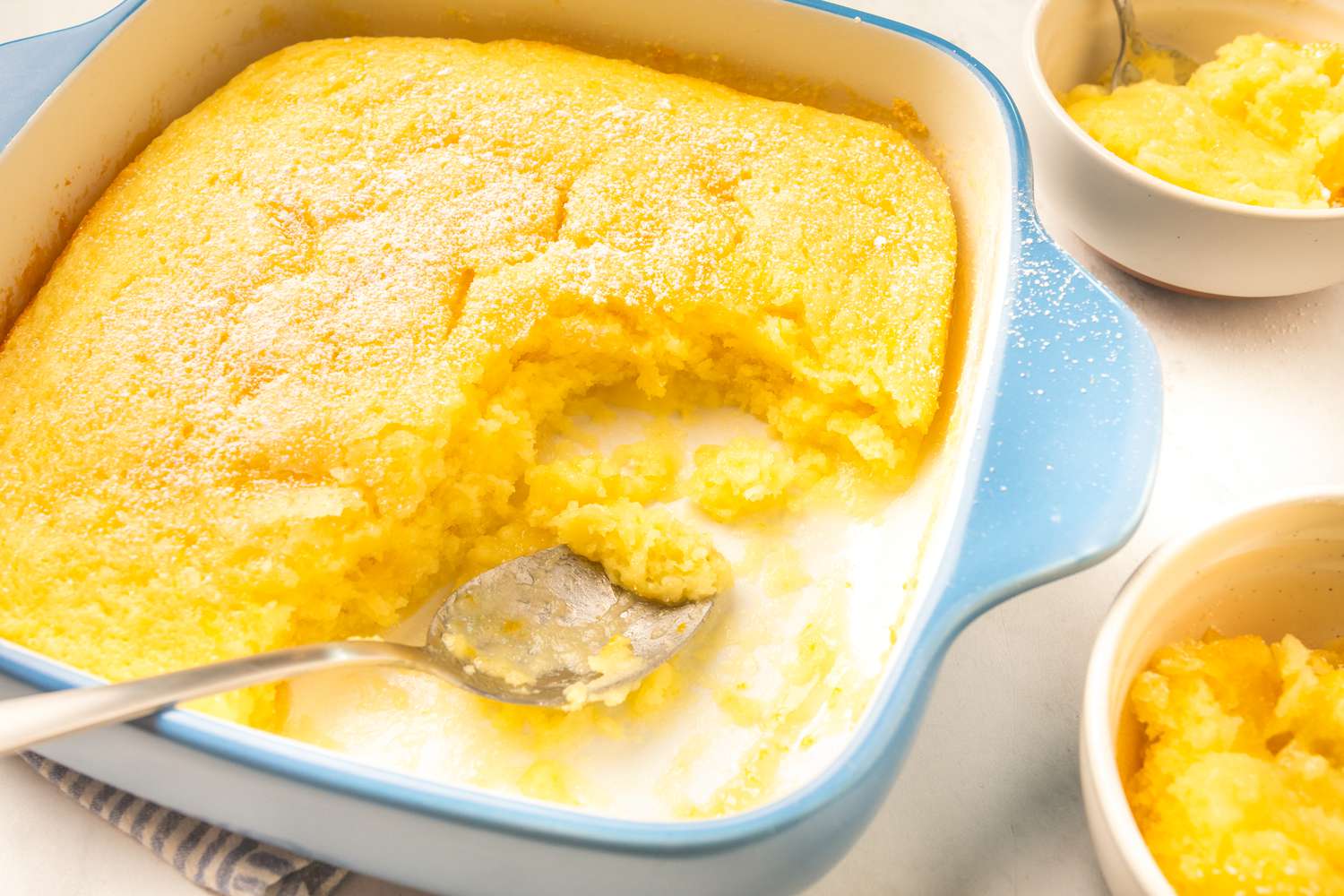 Side angle view of a baking dish with lemon pudding cake with some of the cake scooped out in two small bowls