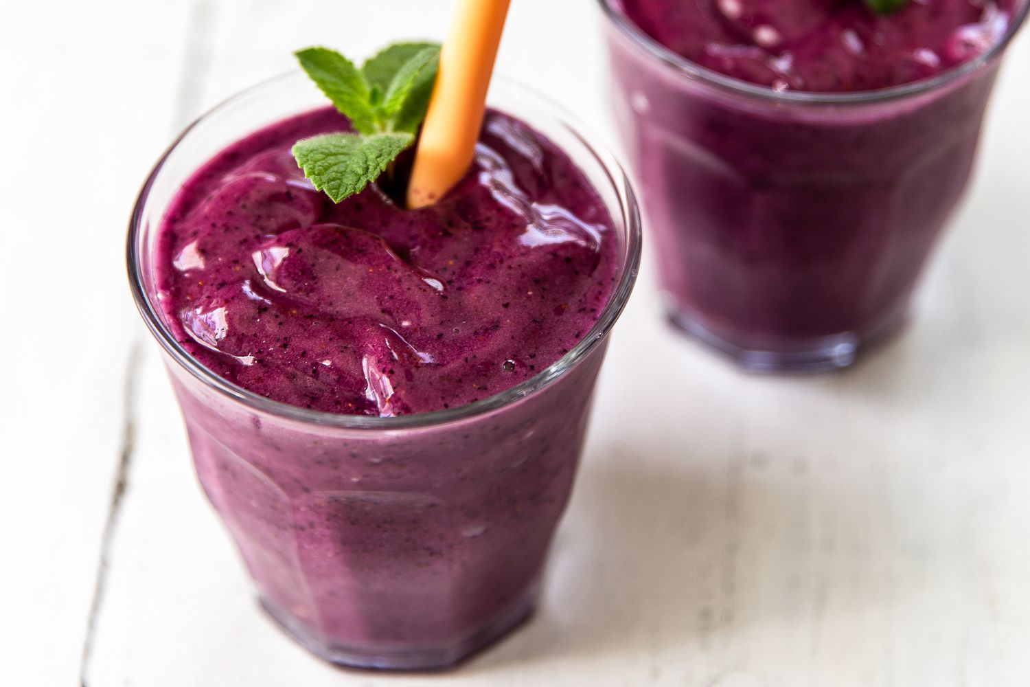 Two glasses of a purple smoothie with mint garnish and an orange straw