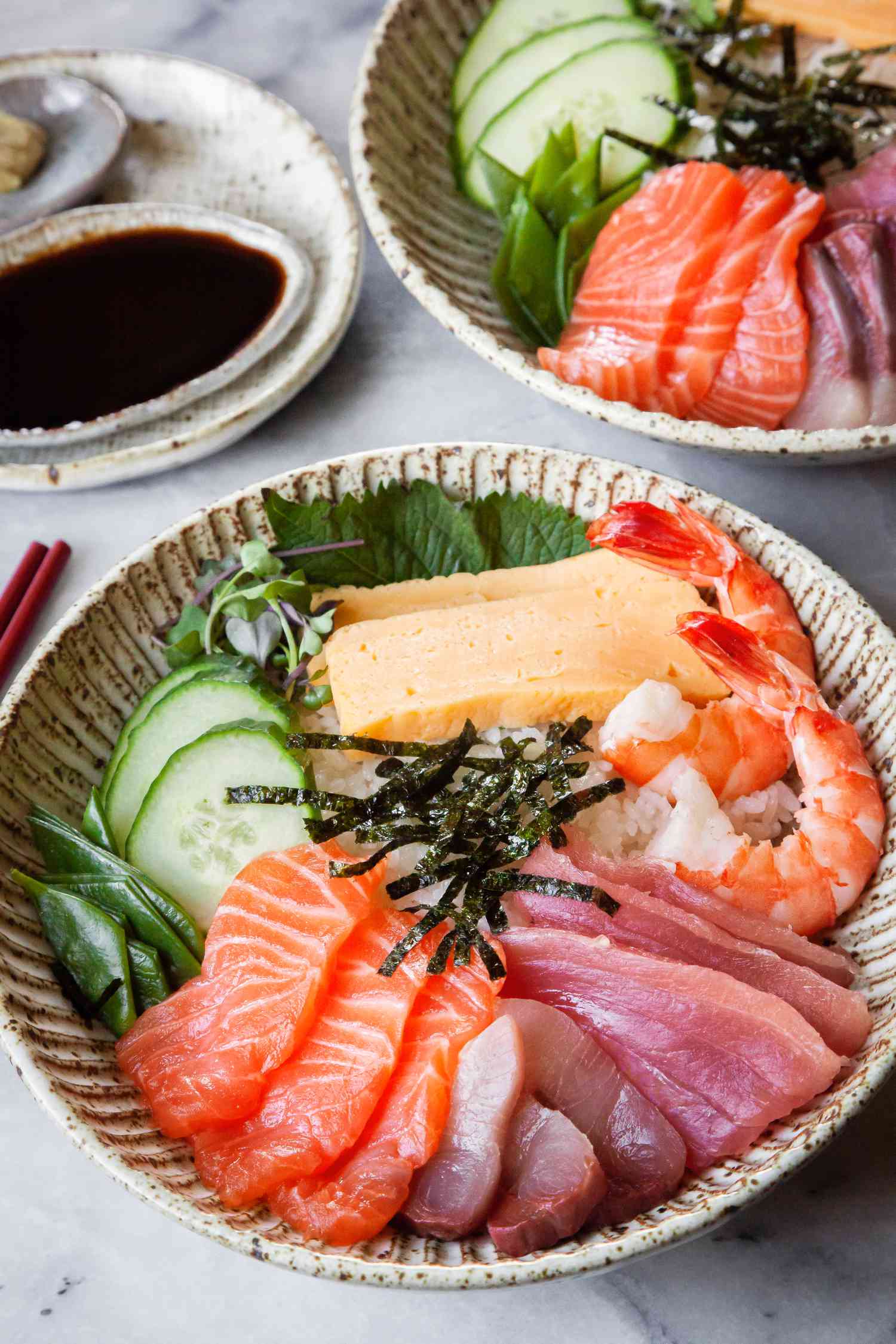 Two Chirashi Bowls with Salmon, Tuna, Shrimp, Tamagoyaki, and Sliced Cucumbers Next to a Small Saucer of Wasabi and Another Small Saucer of Soy Sauce