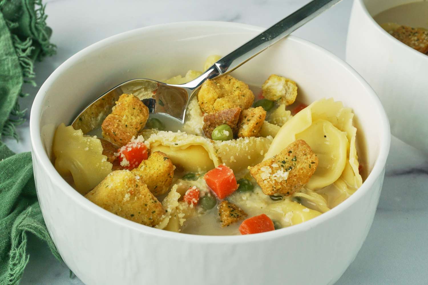 A bowl of creamy tortellini soup with croutons and vegetables featuring a spoon