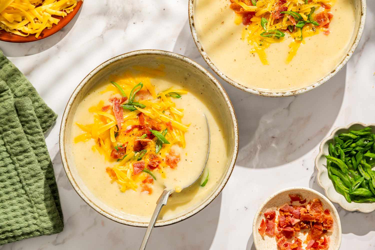 Overhead view of two bowls of quick and easy potato soup topped with bacon, shredded cheese and green onions next to a bowl of bacon and green onions
