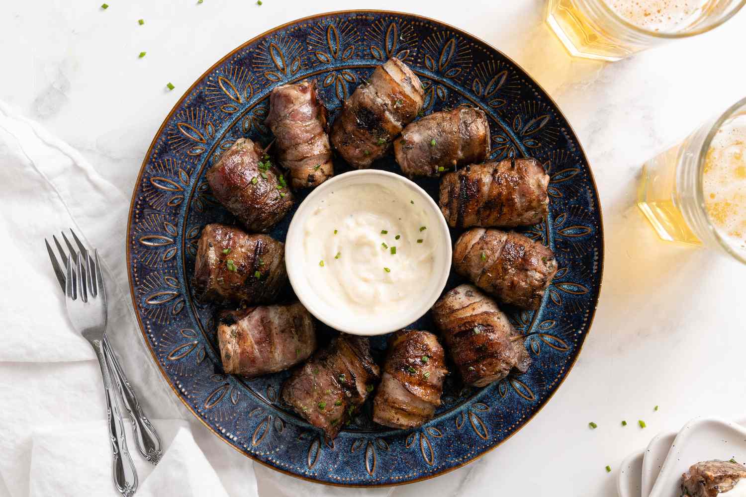 Grilled Bacon-Wrapped Salmon Bites with a Bowl of Lemon Garlic Mayo on a Plate, Surrounded by a Bowl, Spoon, and Glass for Grilled Bacon-Wrapped Salmon Bites with Lemon Garlic Mayo 