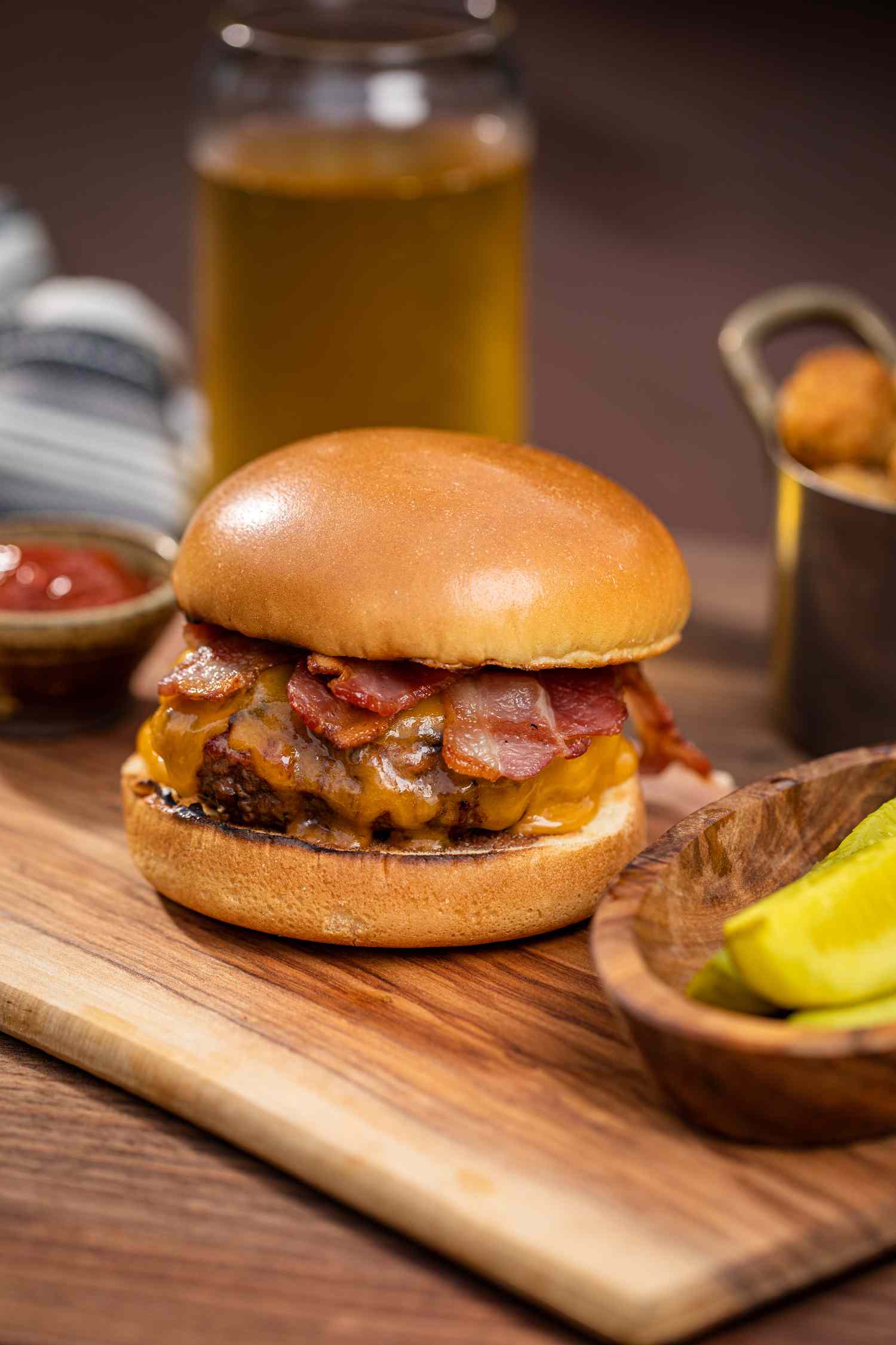 Classic bacon cheeseburger on a cutting board with condiments and drinks around it.