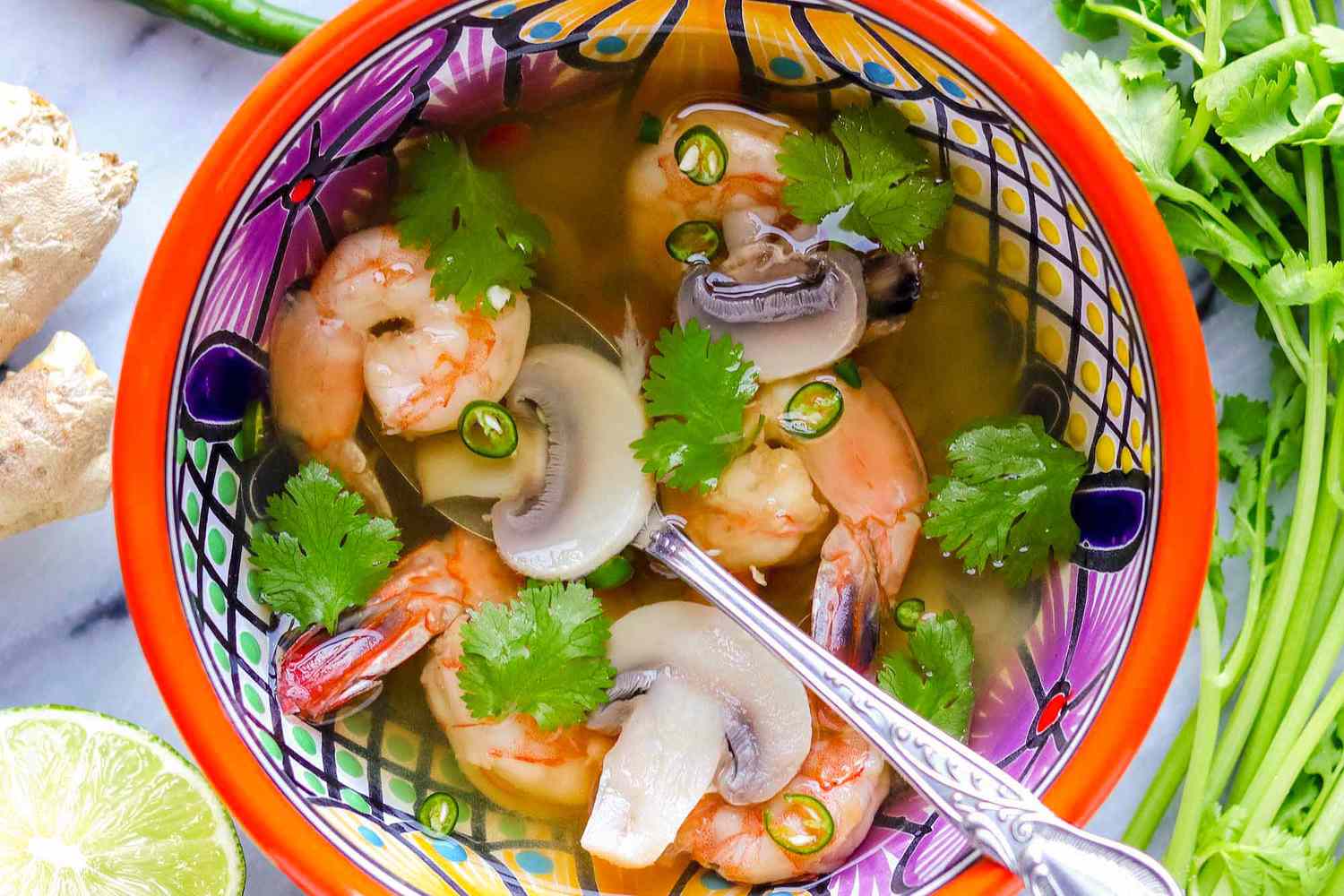 A bowl of tom yum soup with a spoon in it and various ingredients around it