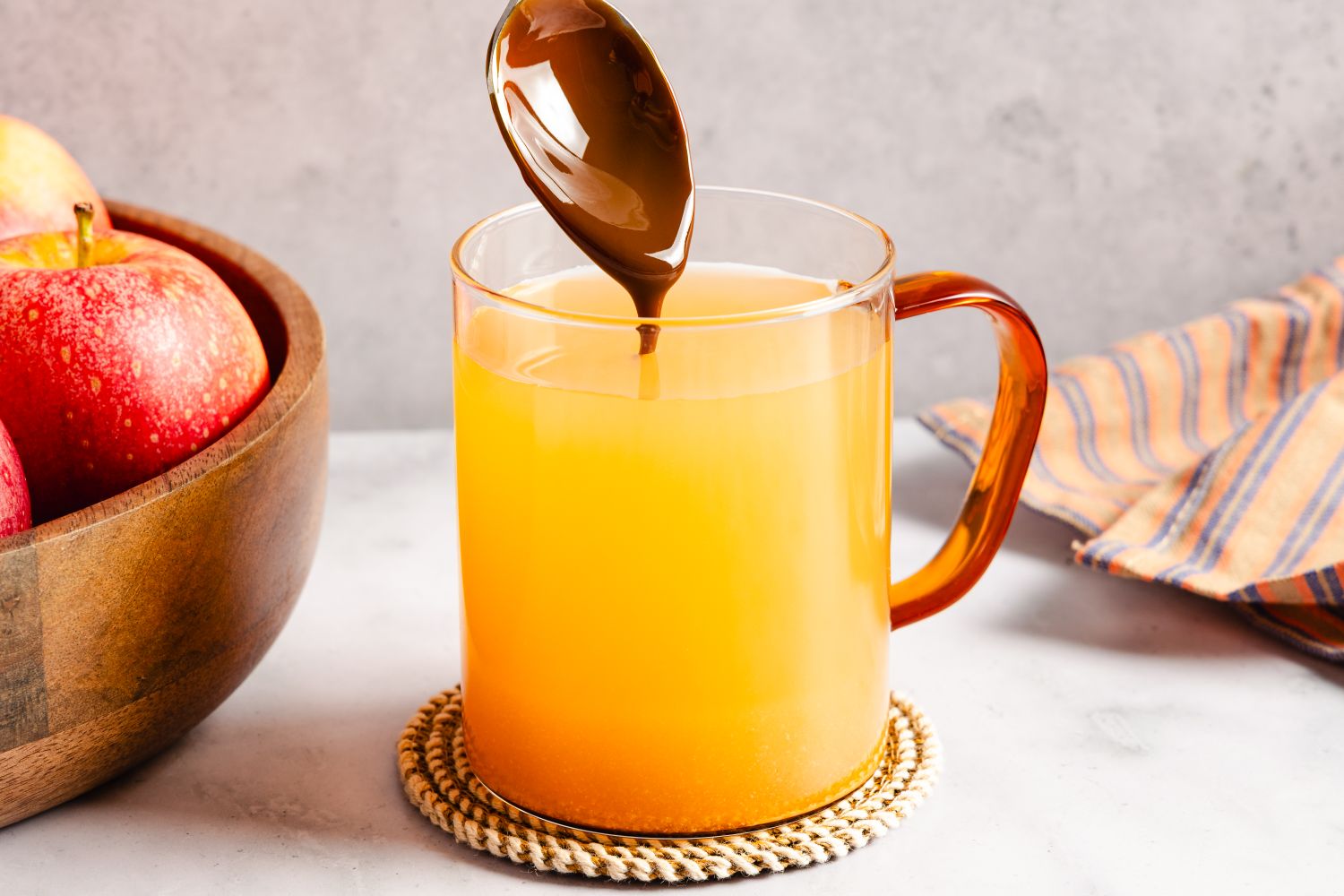 A glass mug filled with apple cider a spoon is pouring caramel sauce into the mug a wooden bowl with apples is nearby