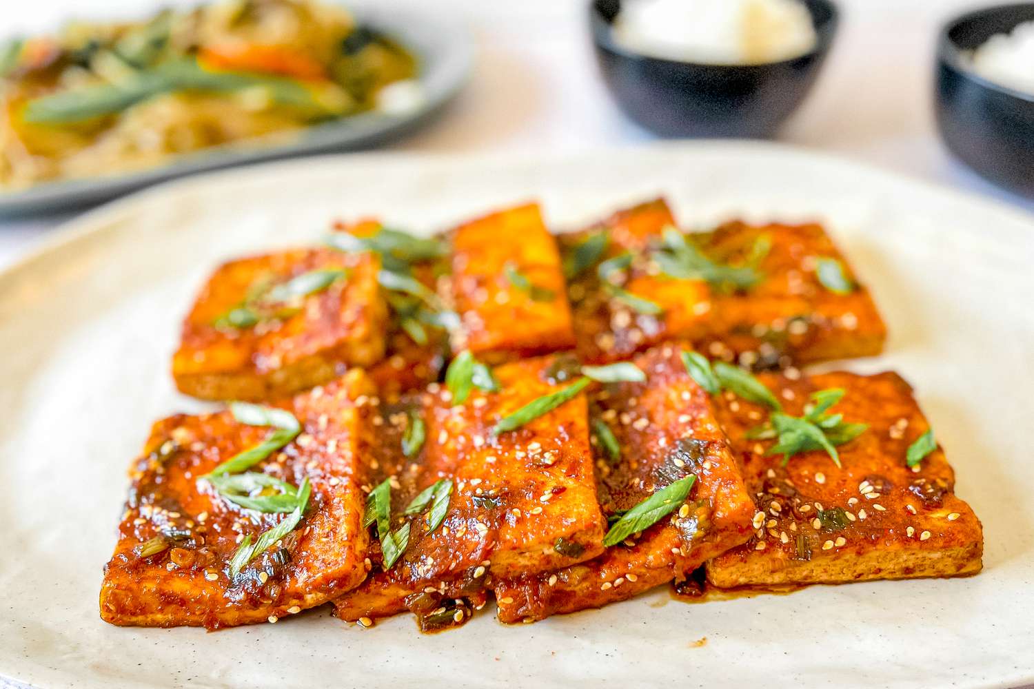 Dubu Jorim (Korean Braised Tofu) on a Platter with Bowls of Rice and Japchae in the Background