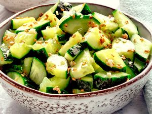 A bowl of sliced cucumbers mixed with sesame seeds, placed on a light-colored surface