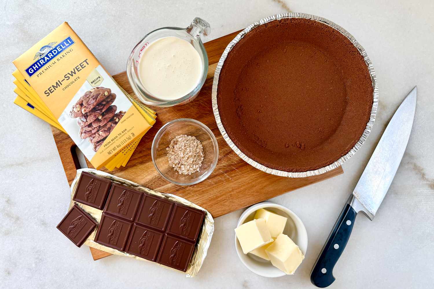 Ingredients for a chocolate tart on a wooden cutting board, including chocolate bars, butter, cream, and a pie crust