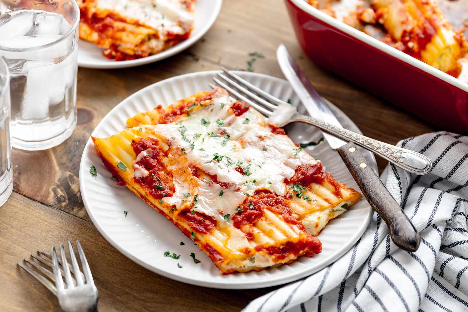 Serving of Manicotti on a Plate With Silverware, and in the Surroundings, a Table Setting With Glasses of Ice Water, a Table Napkin, Forks, Another Plate With a Serving of Manicotti, and a Casserole Dish With More Manicotti