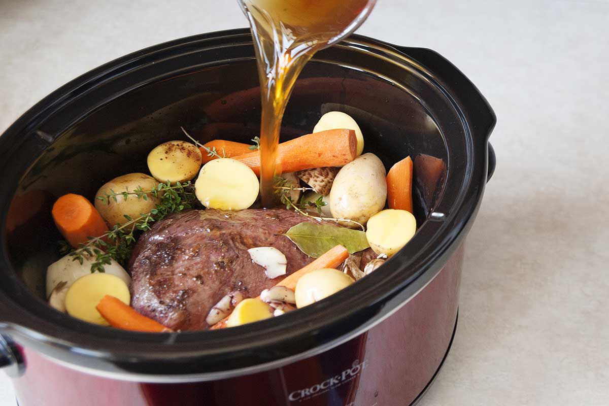Pouring stock into a slow cooker filled with roast, carrots, potatoes and herbsto for the best slow cooker pot roast.
