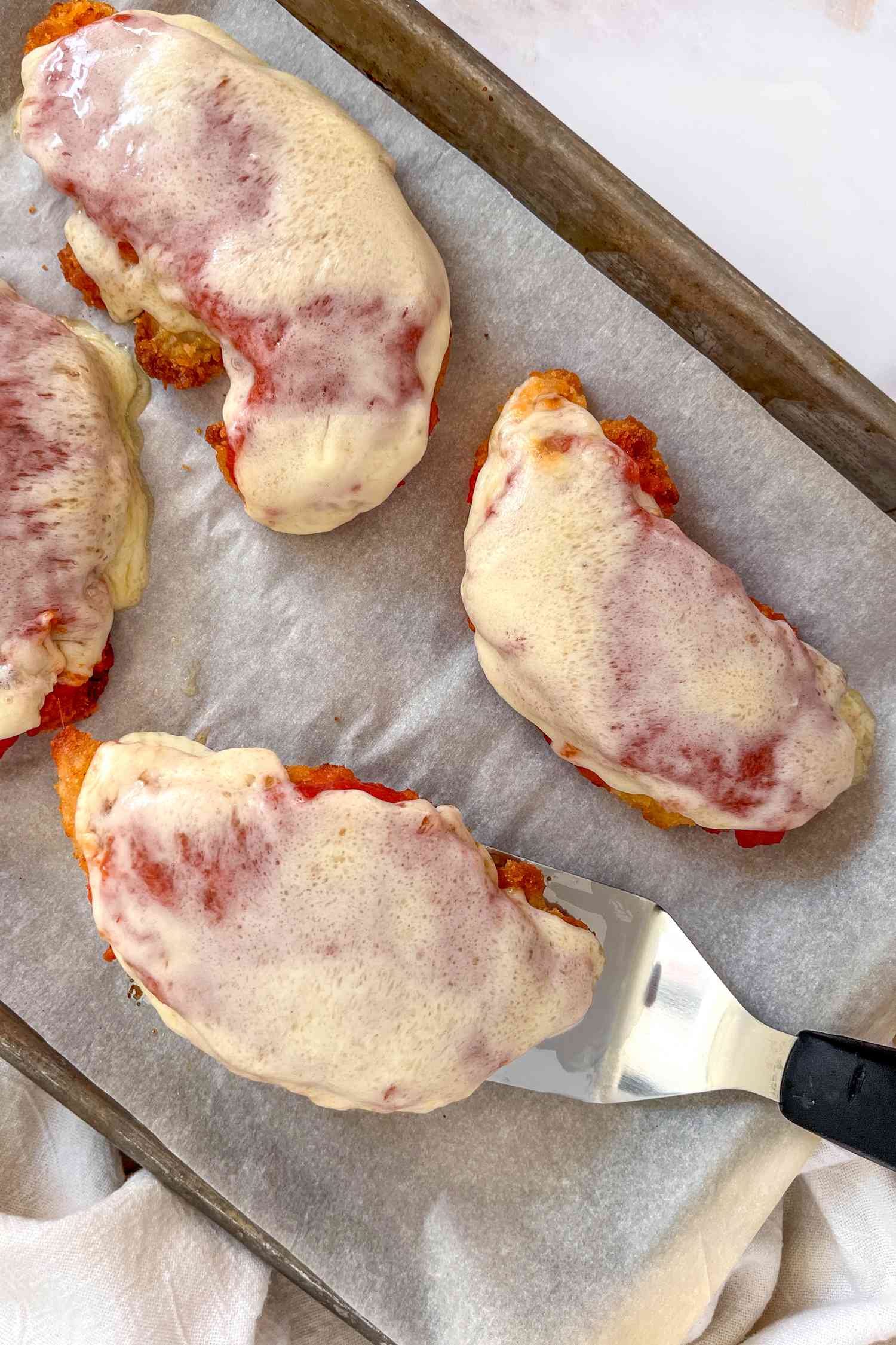Costco chicken parmesan on a parchment paper lined baking sheet with one chicken piece lifted using spatula