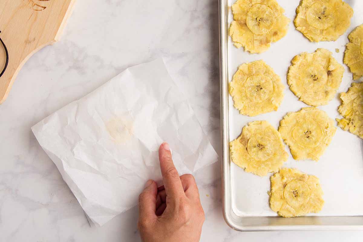 Smashing plantains (tostones) between two pieces of parchment with a coffee mug and setting them on a parchment lined baking sheet.