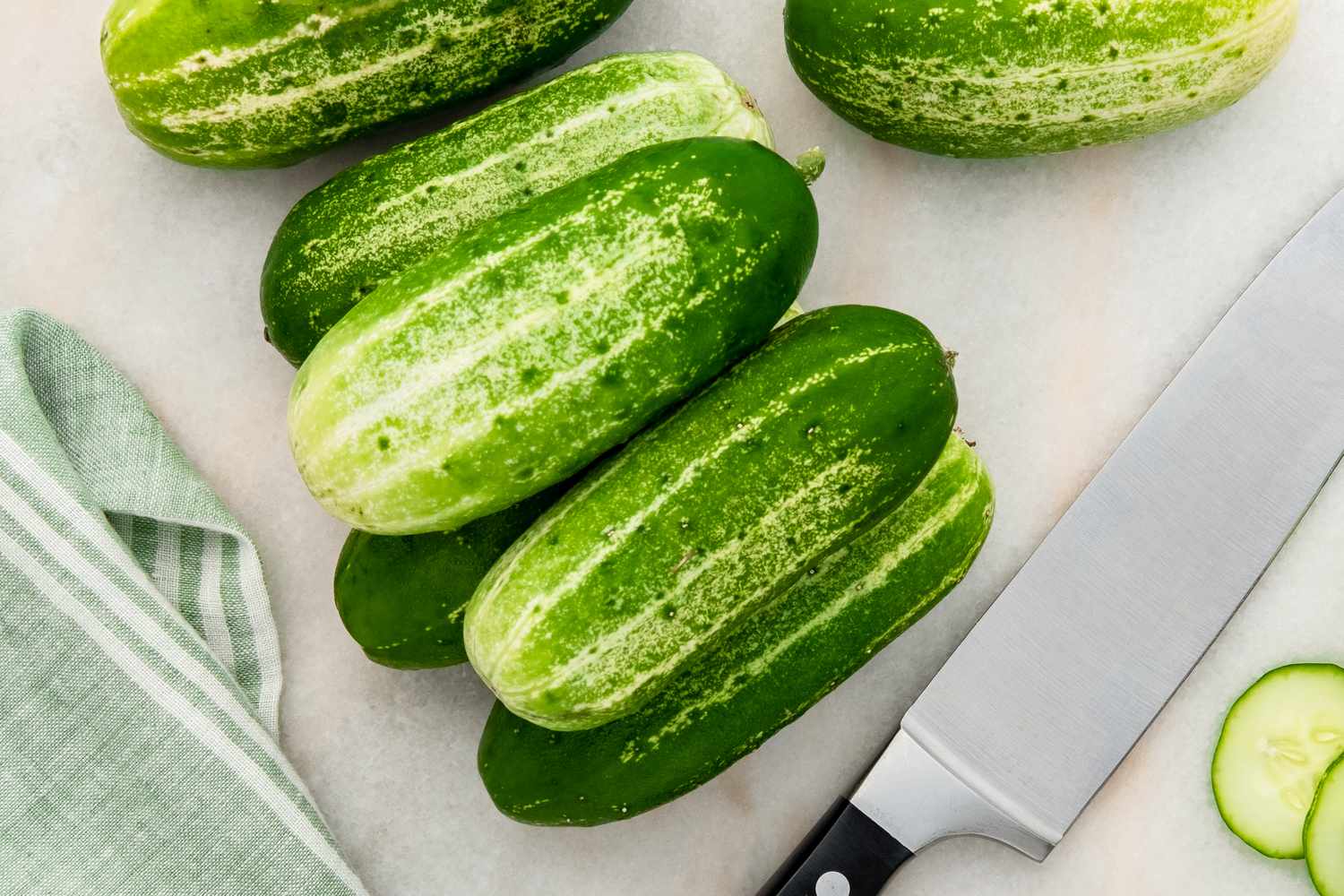 Fresh cucumbers stacked on a kitchen surface with a knife and towel nearby