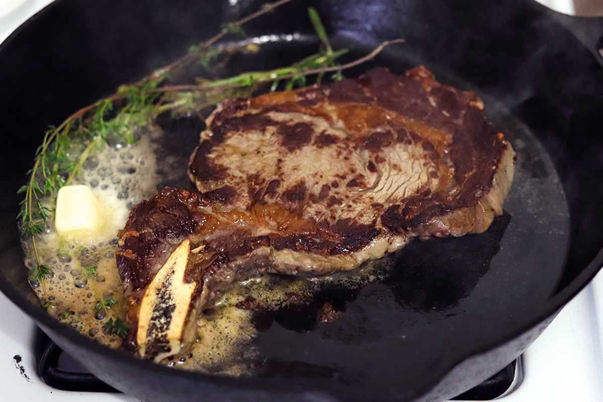 A cast iron skillet with steak cooking inside. Fresh oregano is on the side of the pan and butter is melting overtop. The steak has a dark crust and bone.