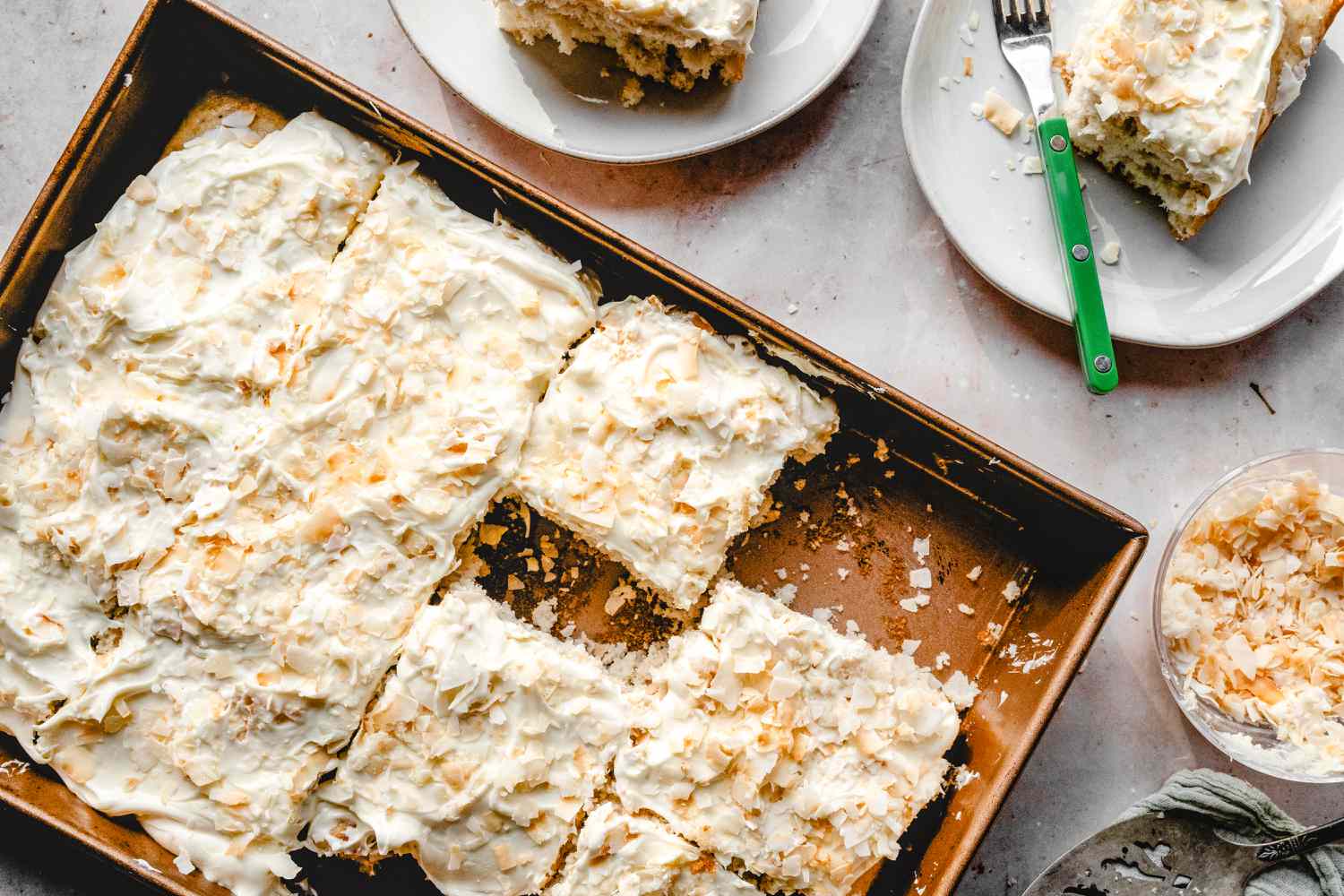 Overhead view of a brown metal baking pan of coconut cake squares surrounded by plates of cake and a bowl of toasted coconut