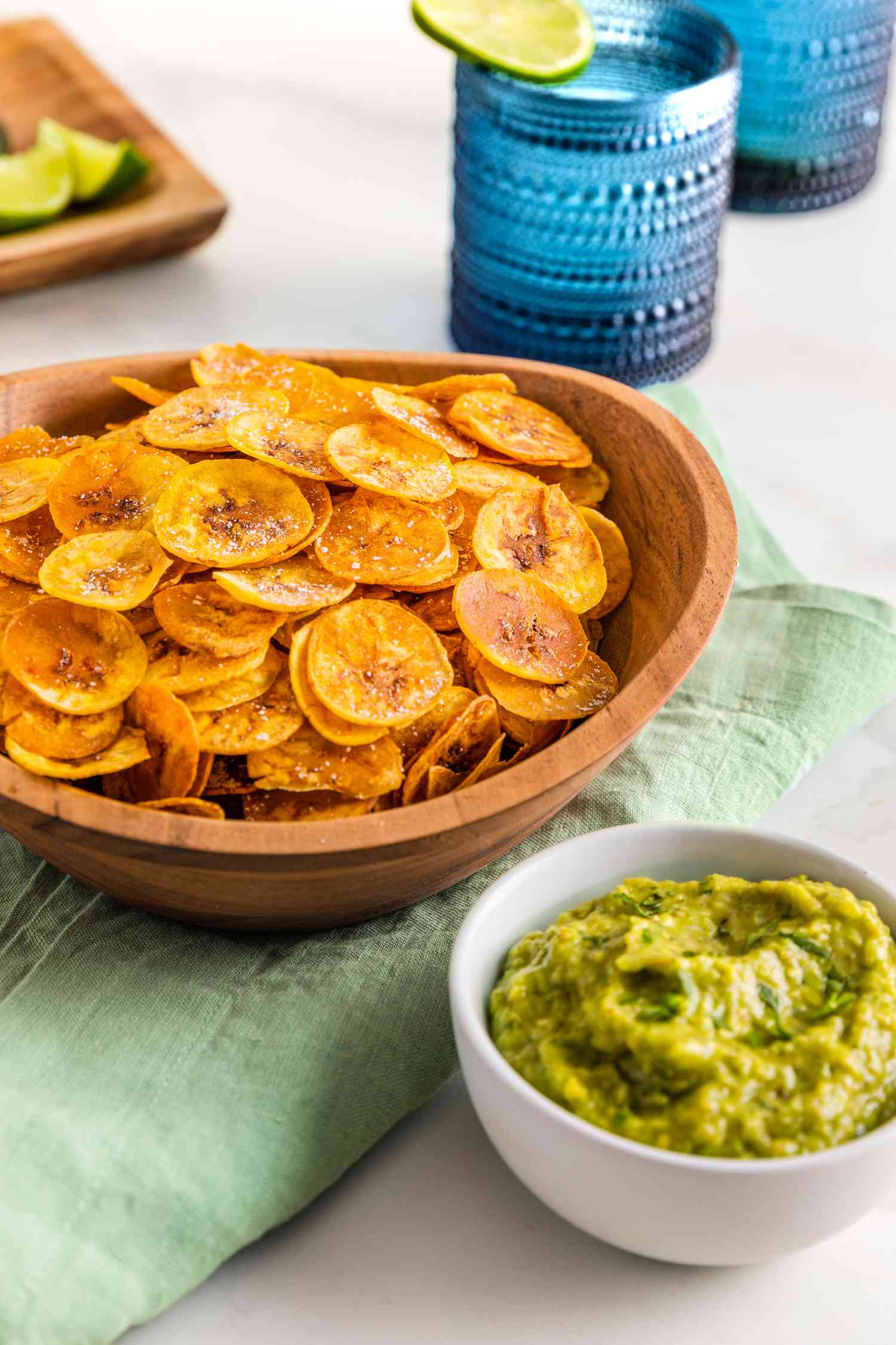 Large Bowl of Plantain Chips, and in the Surroundings, a Bowl of Guacamole, Two Blue Glasses With a Lime Wheel, and a Tray With Lime Wedges