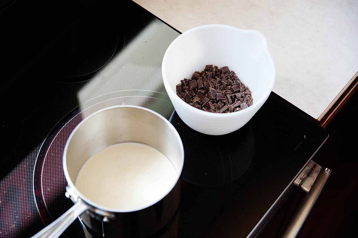 A pan with heavy cream on a stovetop and a white bowl with chopped chocolate next to it for the ganache topping for chocoalte cheesecake.