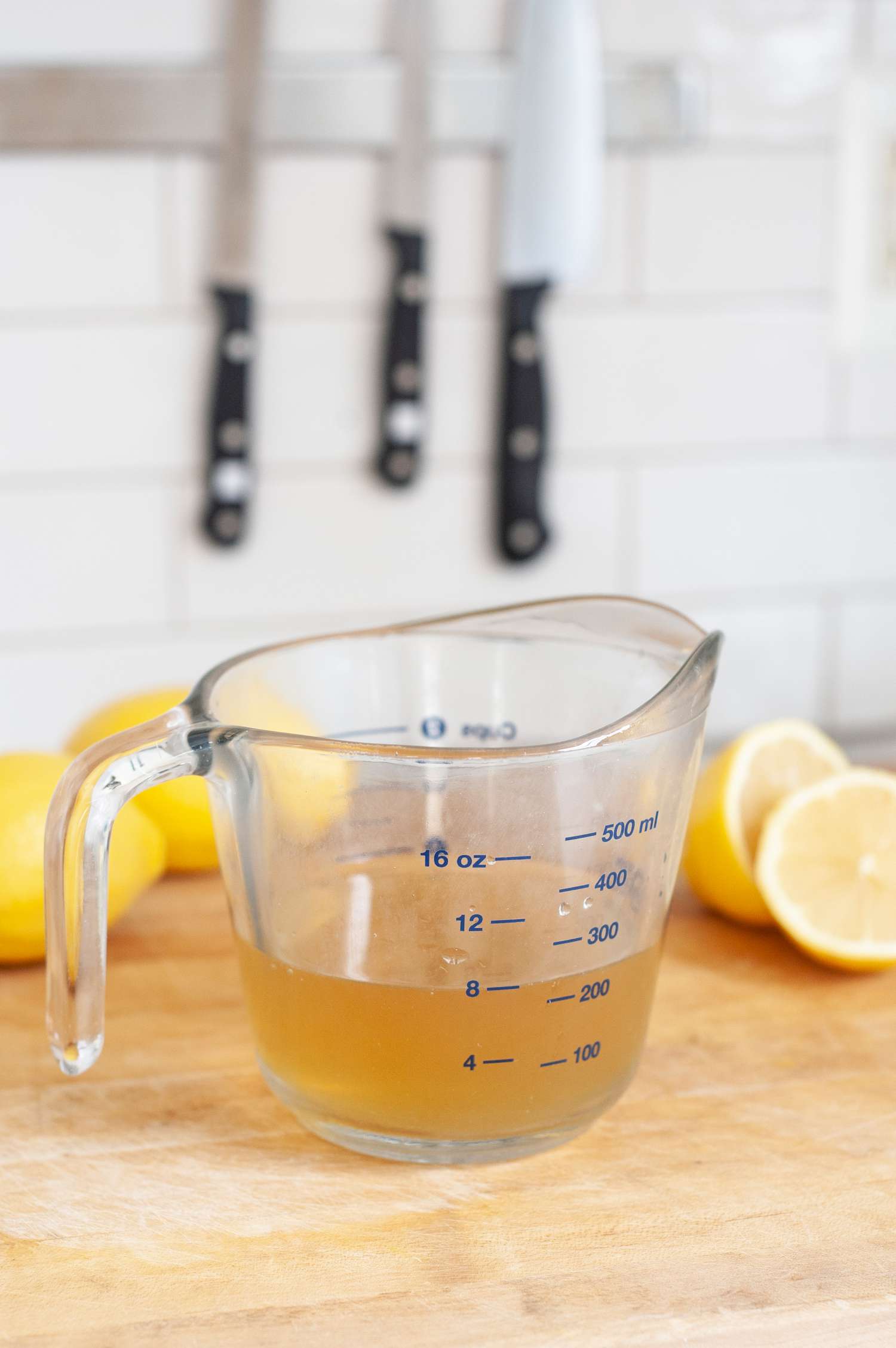 Measuring Cup with 8 Ounces of Broth and in the Background, Lemon Halves, Whole Lemons, and Knives on a Magnetic Knife Holder