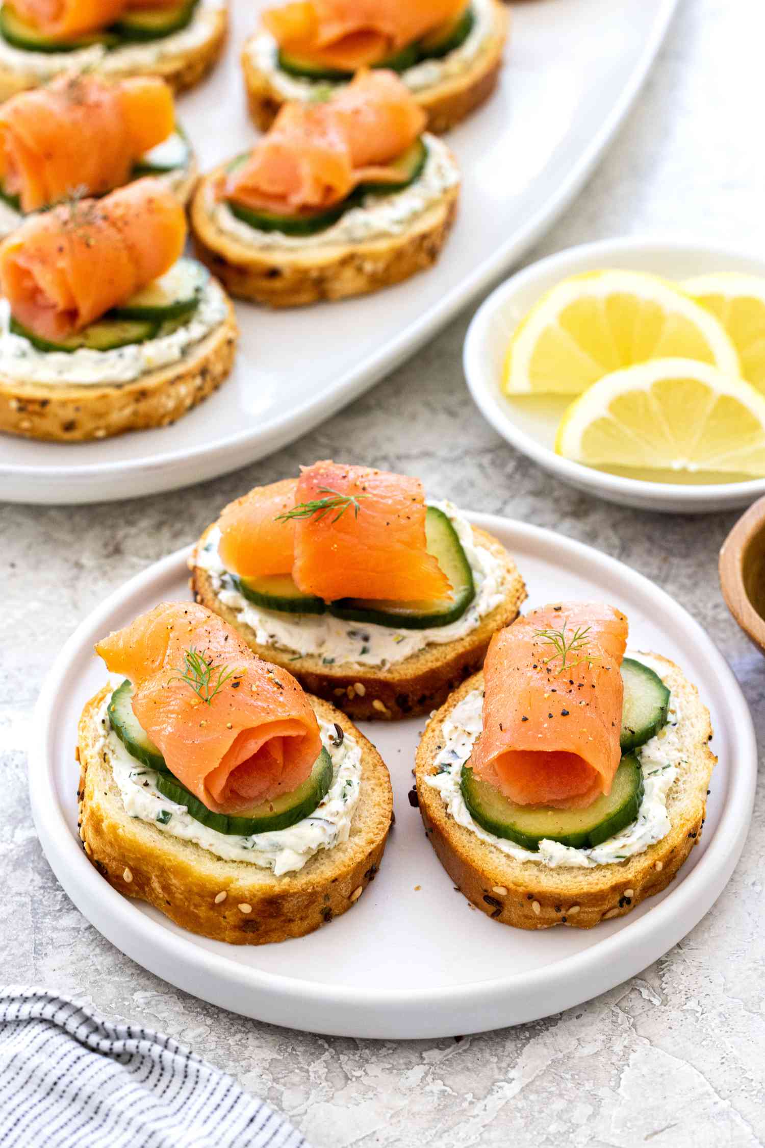 Crostini topped with smoke salmon, pickled cucumbers, and dilled cream cheese on a white plate.
