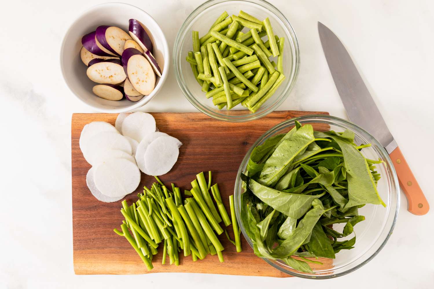 Ingredients for pork sinigang preparation including sliced eggplants green beans radishes water spinach and a knife on a cutting board