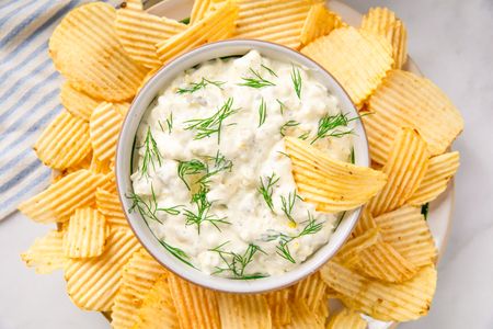 A bowl of pickle dip garnished with dill surrounded by potato chips arranged on a plate