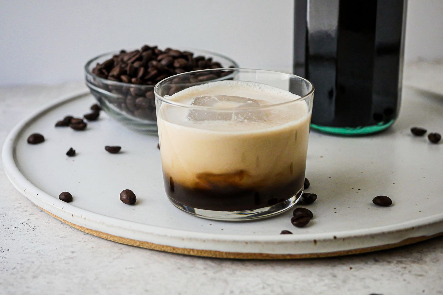 White Russian cocktail on a tray with coffee beans and a bottle of Quick and Easy Vegan Coffee Liqueur.