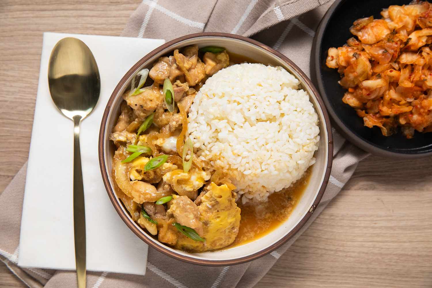 Microwave Oyakodon and Rice in a Small Plate Surrounded by a Table Setting with a Spoon, a Table Napkin, and a Small Plate with Kimchi