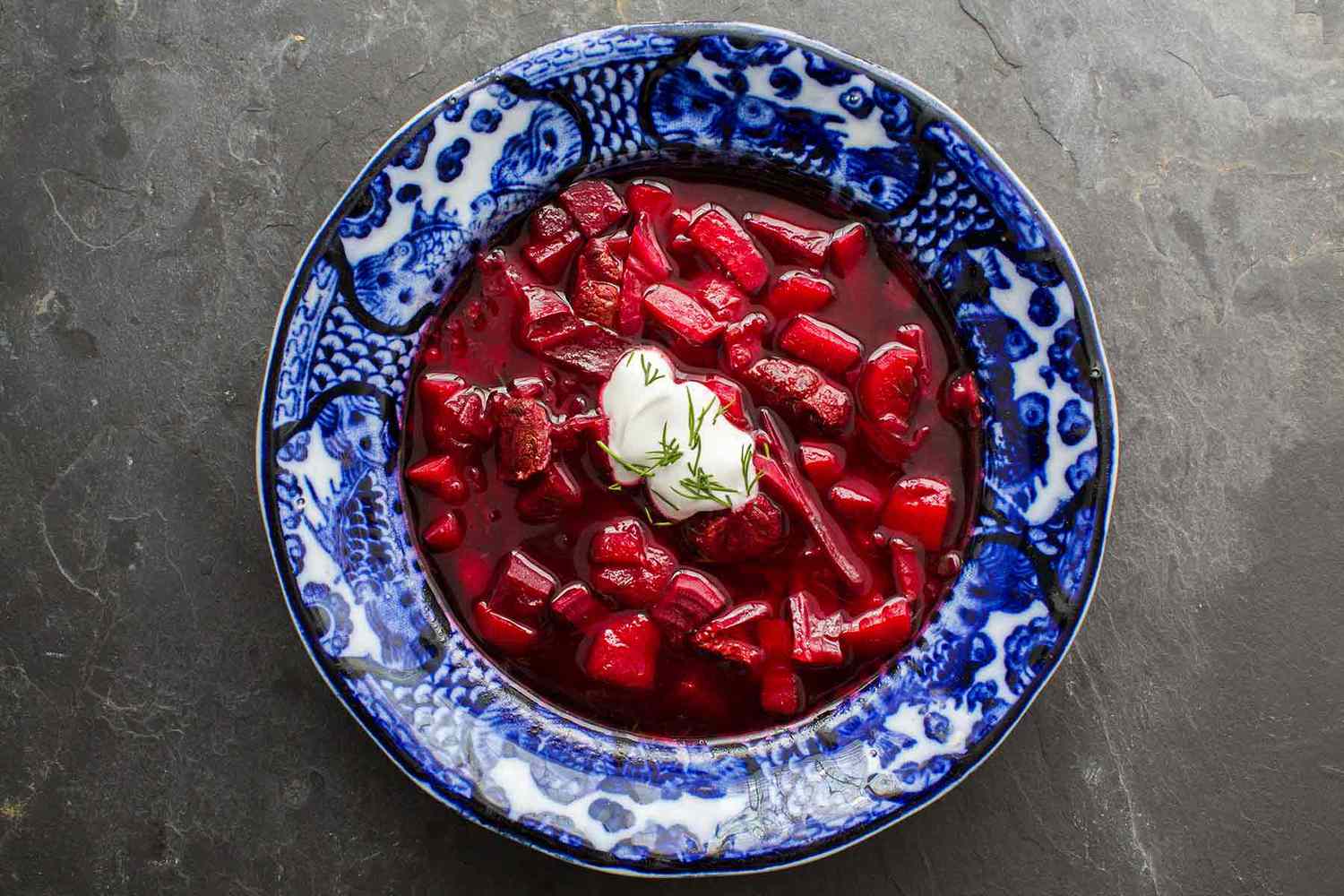 A bright blue bowl on a gray background with borscht and sour cream and dill