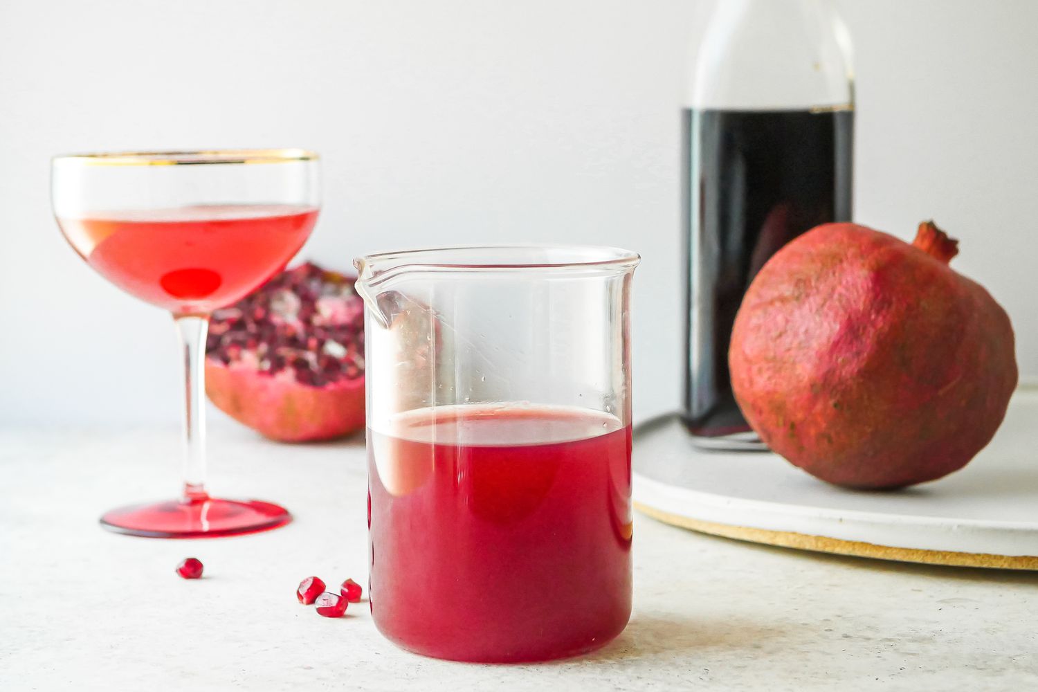 Grenadine Cocktail Syrup in a cocktail mixing glass with a cocktail behind it and pomegranates.