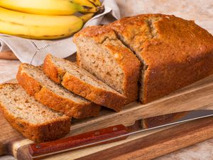 A partly sliced banana bread on a cutting board, with bananas in the background