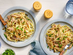 Linguine with tuna and capers on two plates at a table setting with a bowl of parsley, a denim table napkin, salt and pepper shakers, and a glass of water
