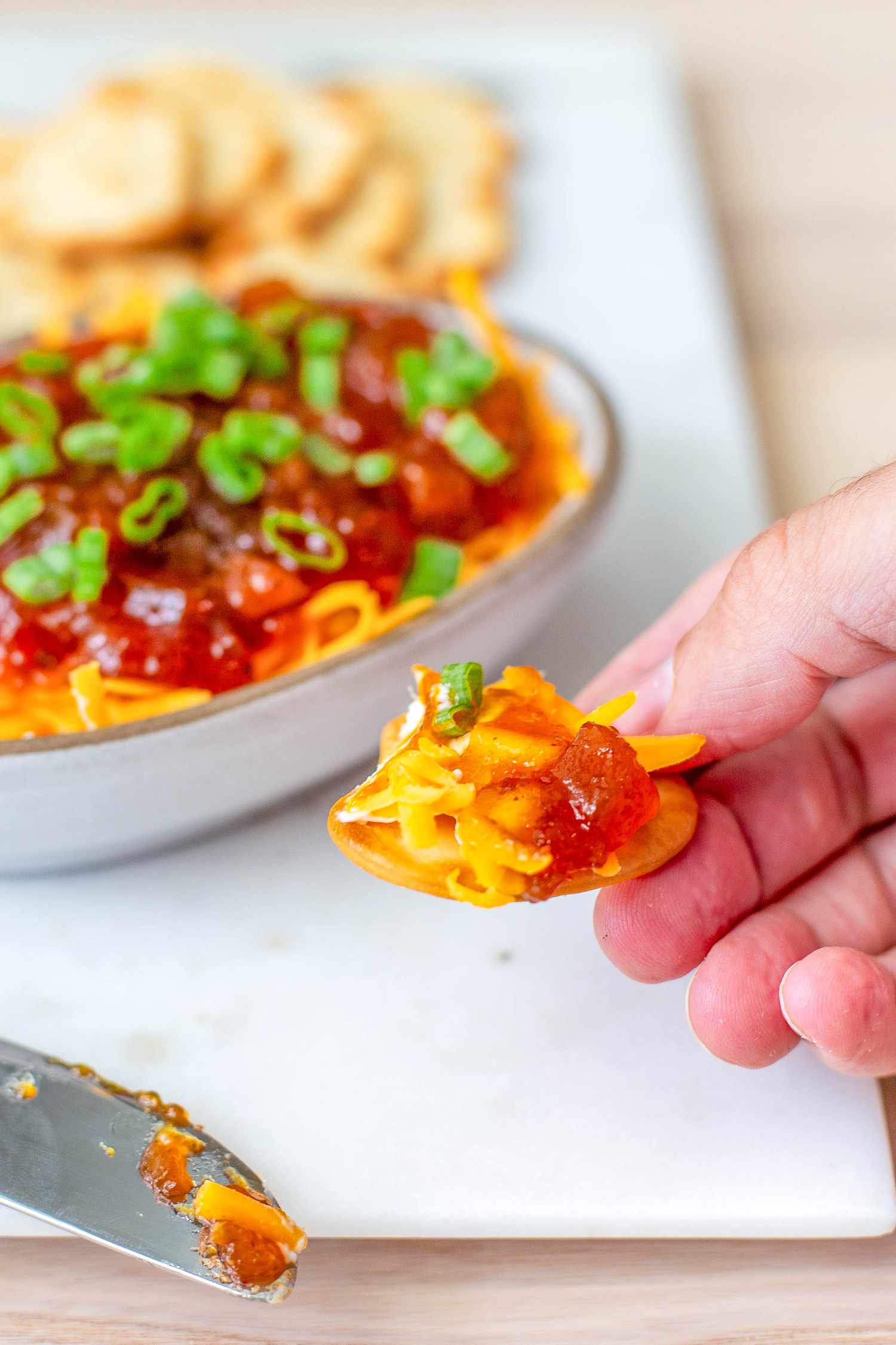 Hand lifting a cracker with chutney cheese appetizer, and in the background, more on the platter 