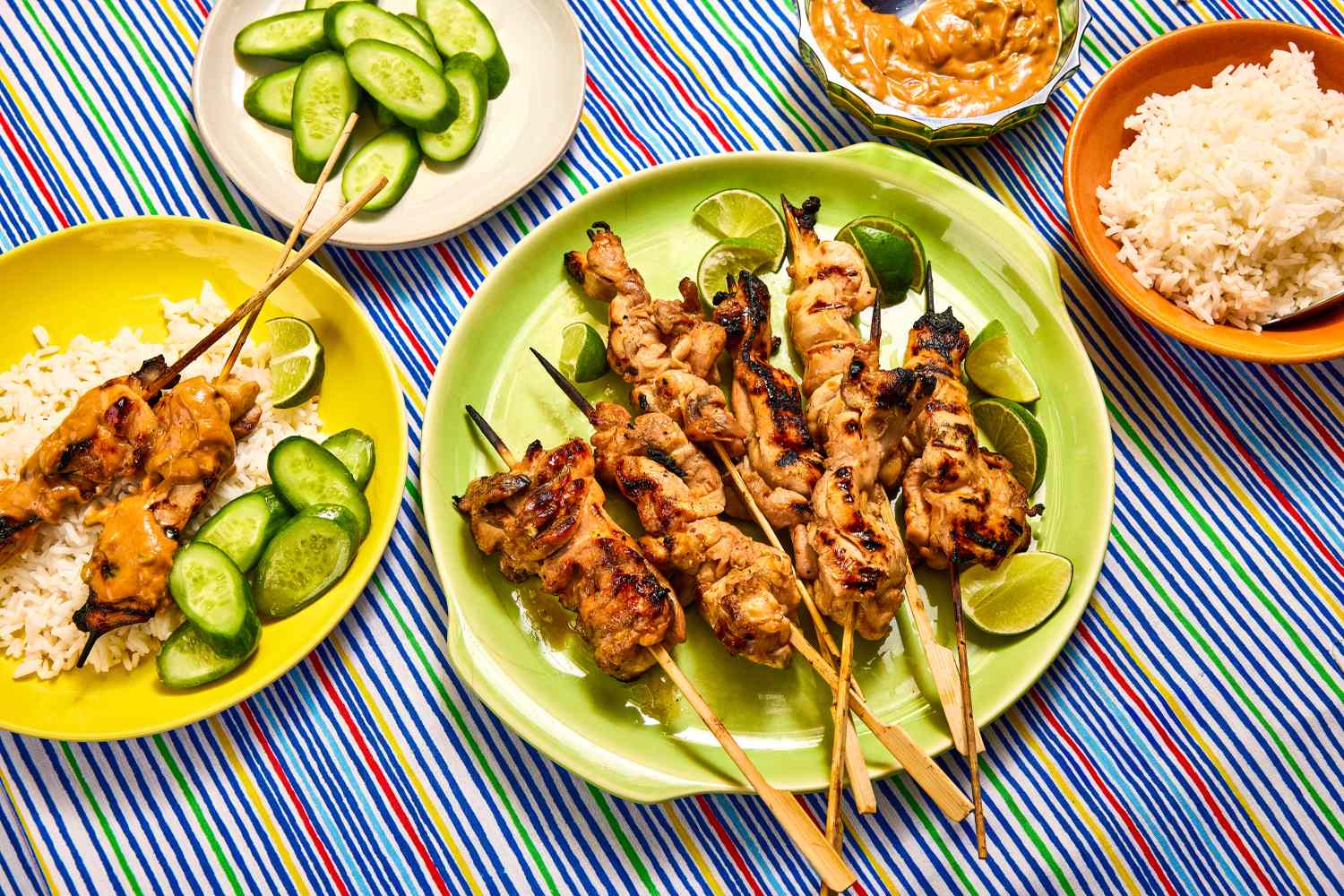 Plates of grilled chicken skewers, cucumber slices, rice, and peanut sauce on a striped tablecloth