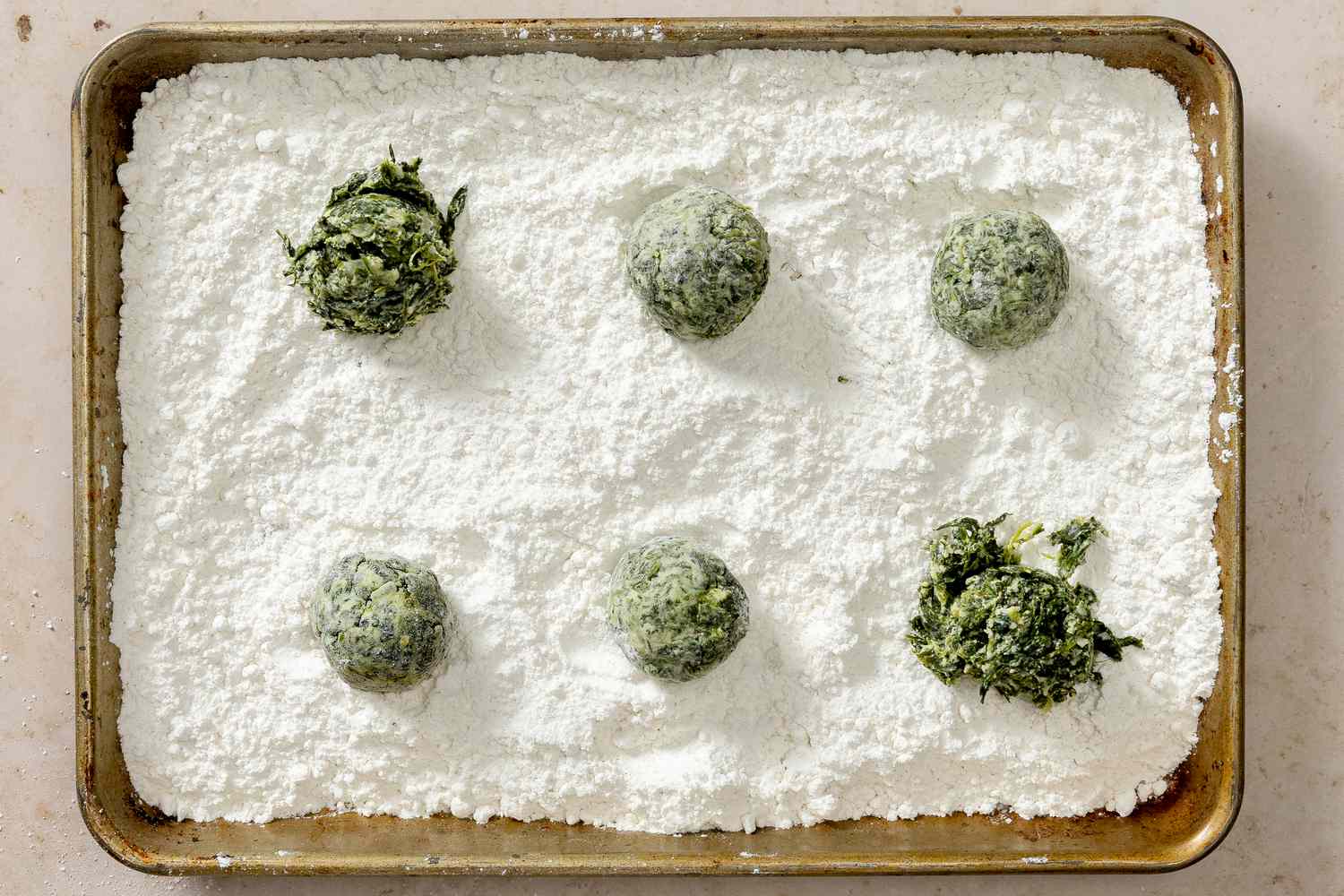 Overhead view of a baking sheet with a layer of flour and some formed and some unformed dumplings of ricotta mixture for Ravioli Gnudi Recipe