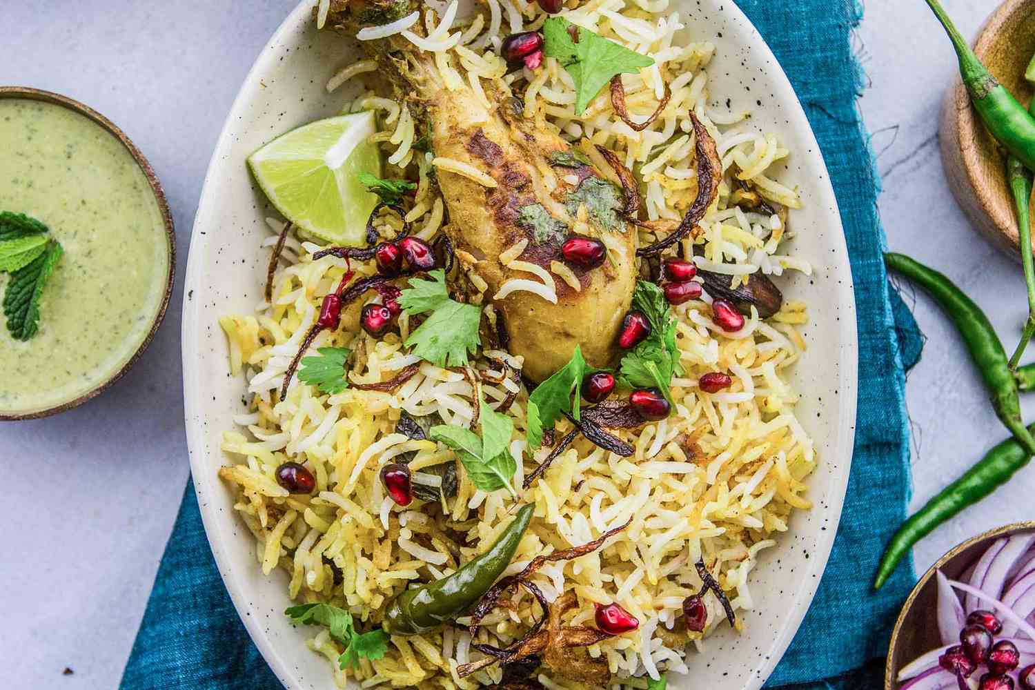 Hyderabadi Chicken Biryani on a Platter Topped With Pomegranate Seeds and a Lime Wedge, and in the Surroundings, a Bowl of Raita, a Bowl With Onion Slices and Pomegranates, and Another Bowl With Chilis Cascading Onto the Counter, All on a Teal Kitchen Linen 