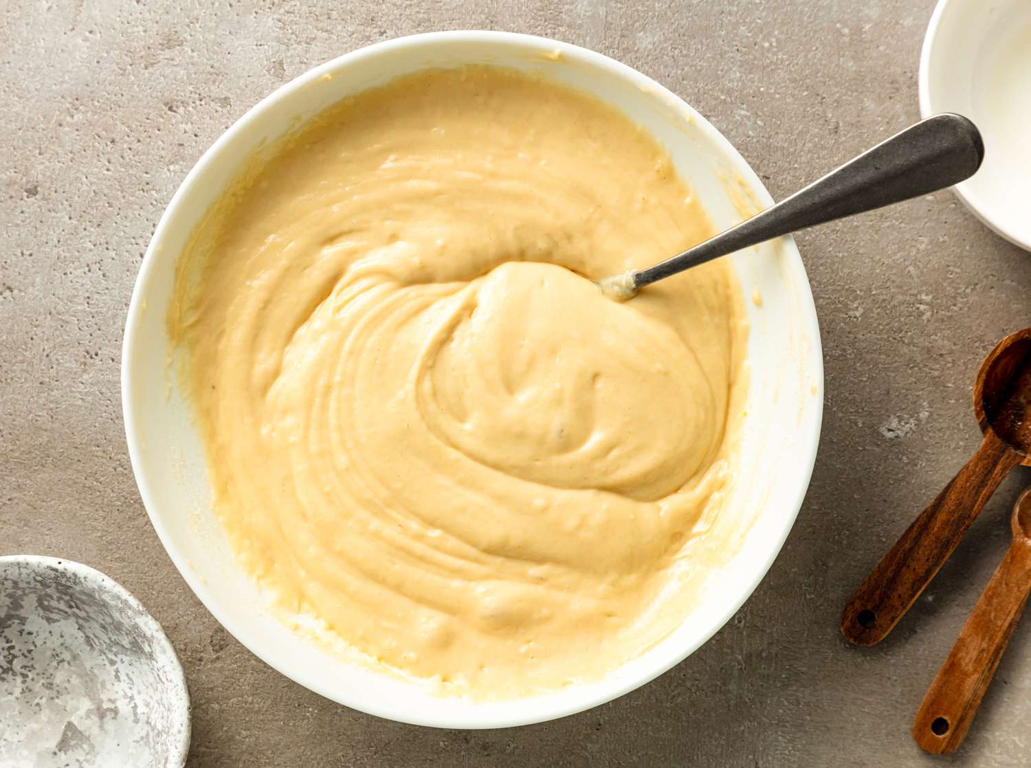 A bowl with pancake batter and a spoon next to measuring spoons and a small empty dish