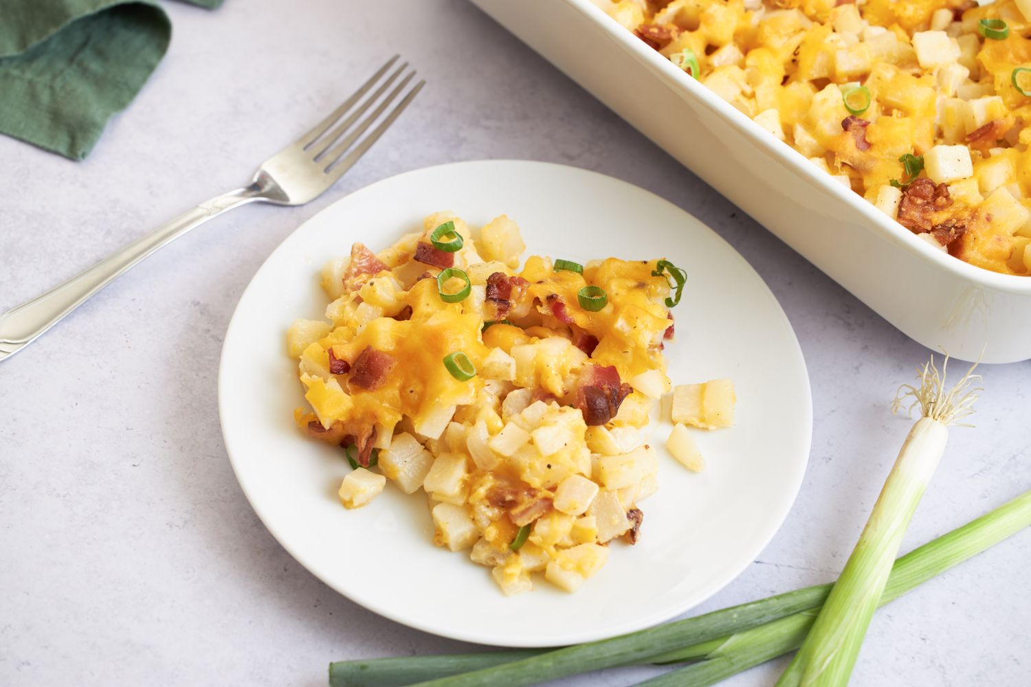 A plate of baked diced potatoes topped with cheese bacon and green onions next to a casserole dish of the same
