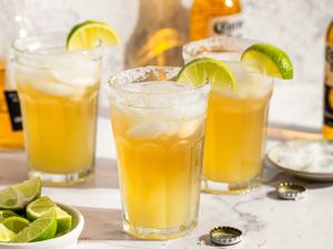 Three glasses of bebidas garnished with lime wedges with bottles and lime wedges in the background
