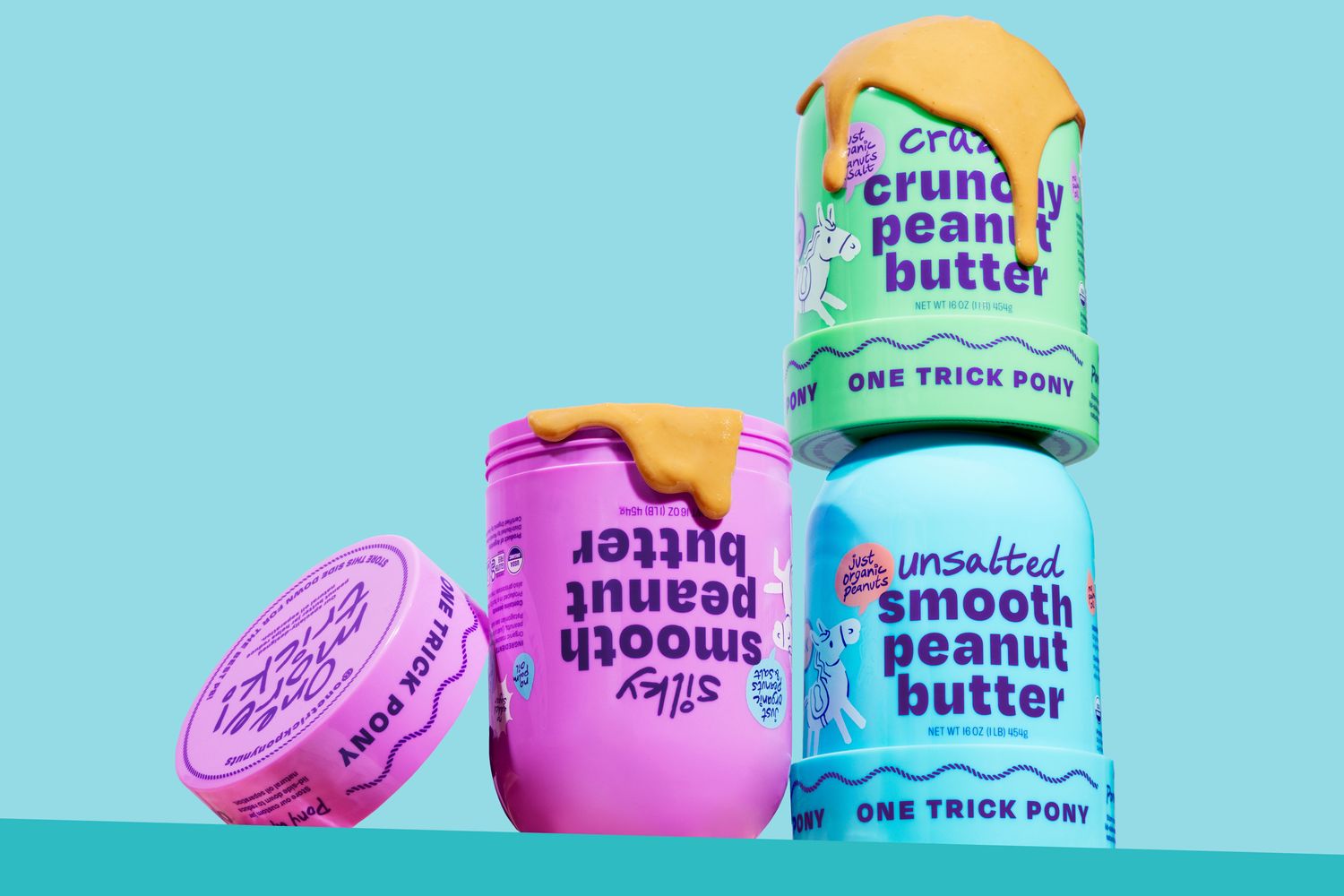 Four jars of peanut butter stacked artistically with one lid off showcasing smooth and crunchy varieties from a brand called One Trick Pony