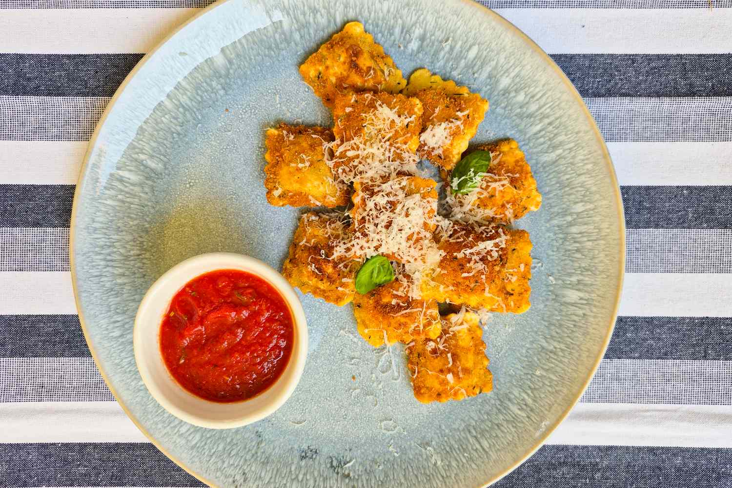 Plate of crispy ravioli with grated cheese and a small bowl of marinara sauce on the side arranged on a striped background