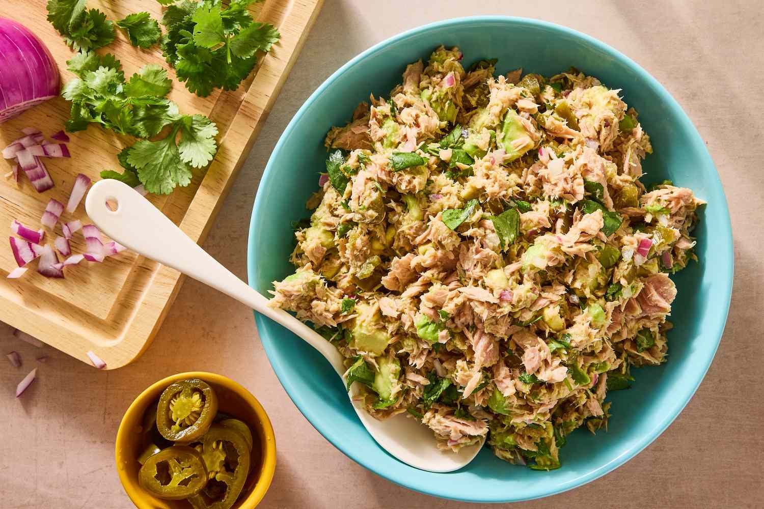 A bowl of tuna and avocado salad with a spoon jalapeos and chopped herbs nearby