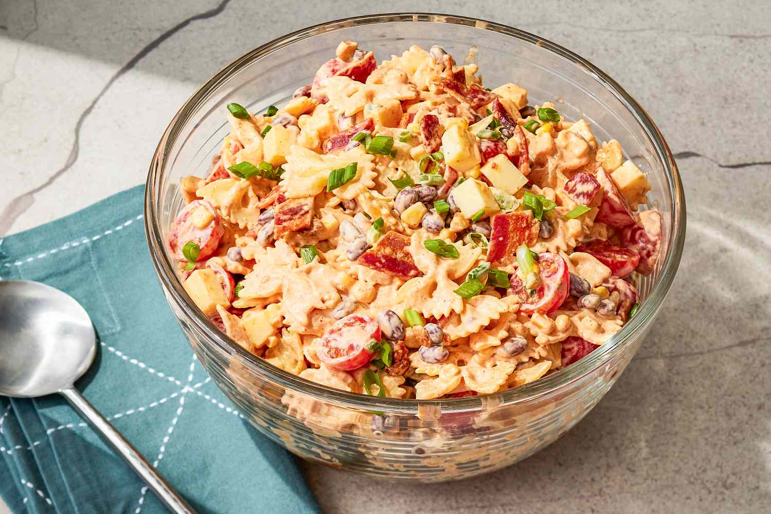A bowl of cowboy pasta salad containing farfalle pasta, diced meats, cheese, and chopped vegetables, next to a blue napkin and spoon
