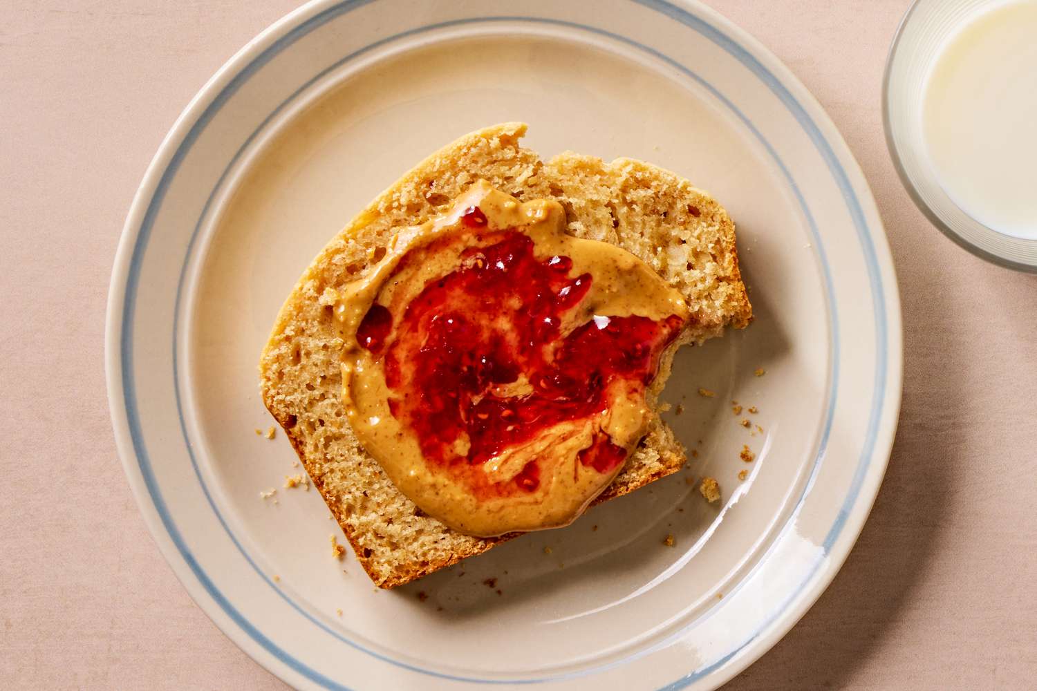 Overhead view of a plate with a slice of retro peanut butter bread topped with peanut butter and jelly next to a glass of milk