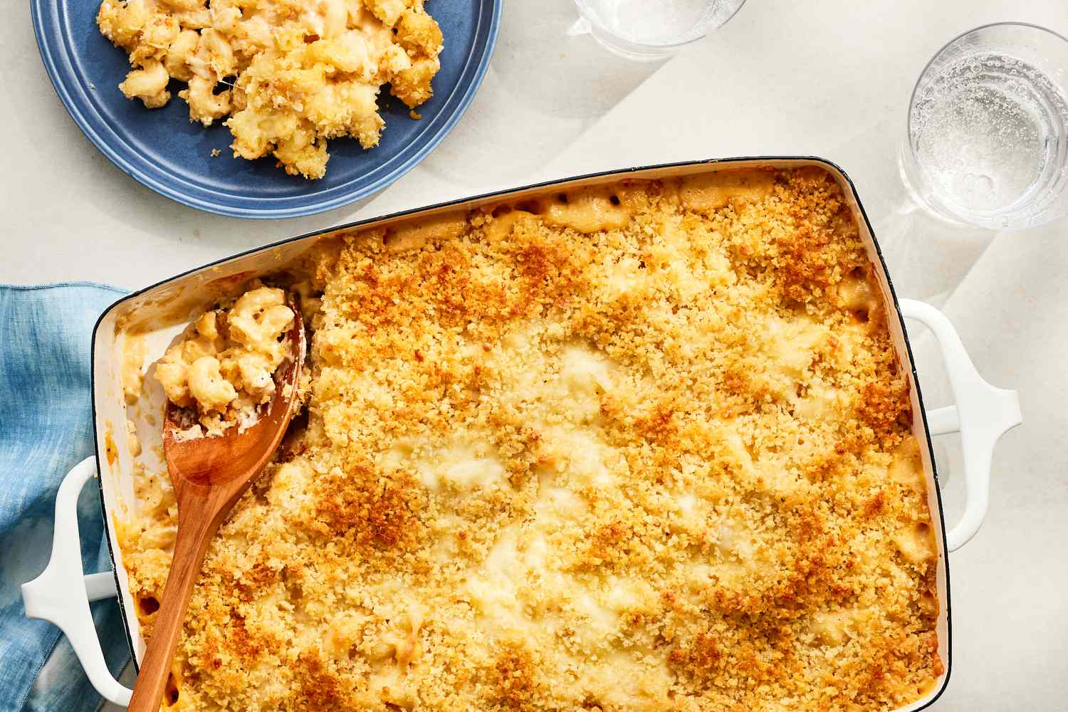 Overhead view of million dollar macaroni and cheese topped with panko in a baking dish with a serving spoon in the corner, all next to a small blue plate with a serving