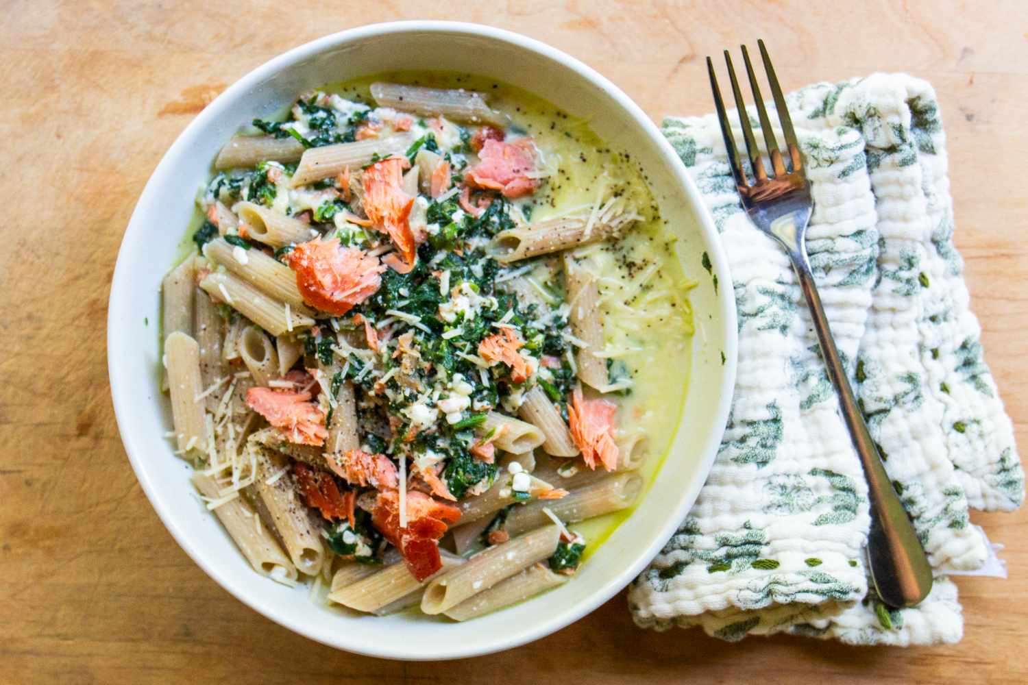 Bowl of pasta with smoked salmon and greens on a wooden table fork and cloth napkin on the side