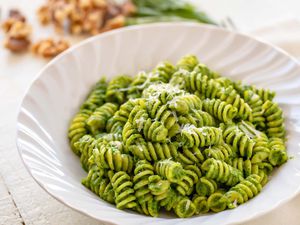 Horizontal view of a white bowl with rotini and kale pesto inside. Walnuts and basil are in the background.
