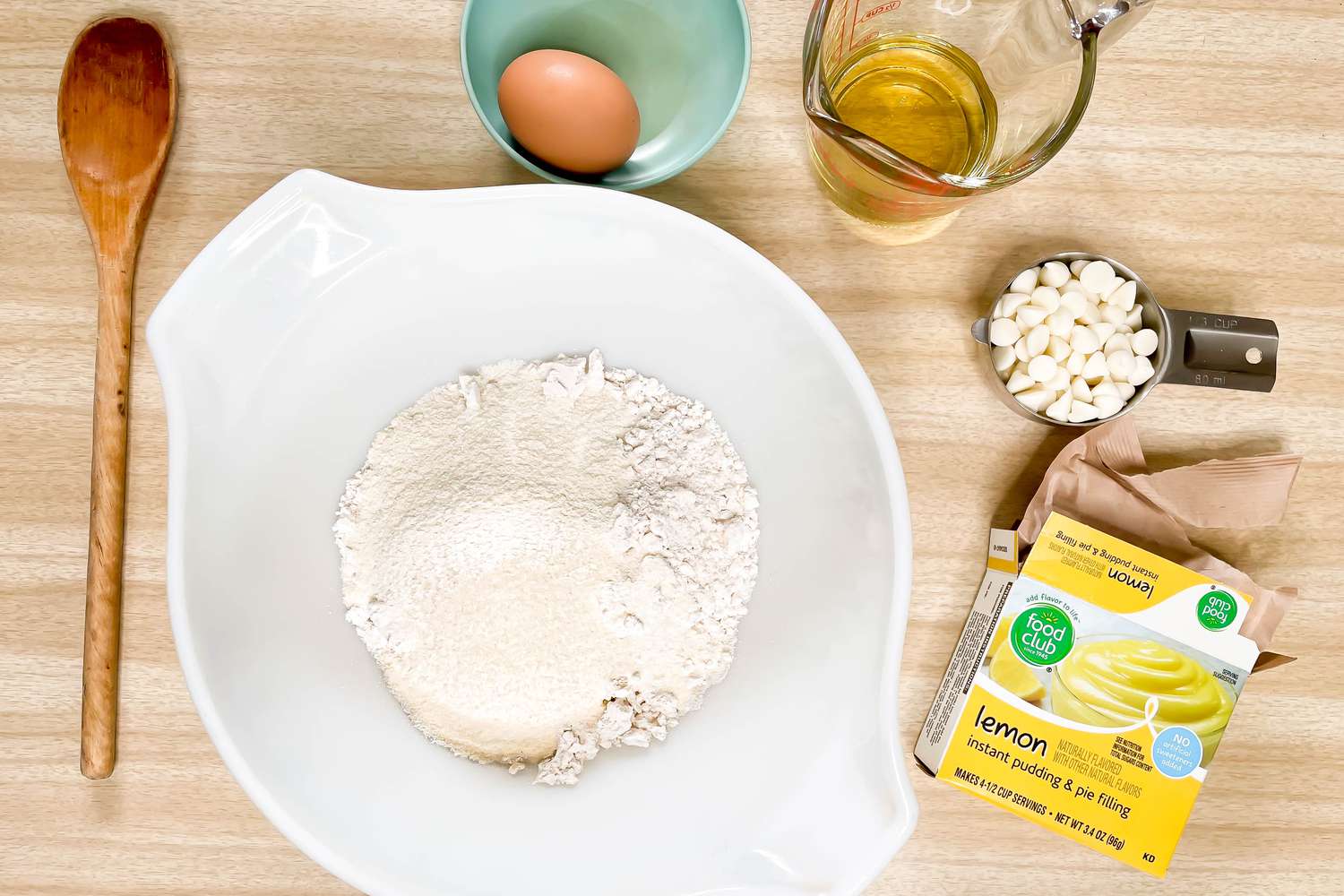 5-Ingredient Pudding Mix Cookies Ingredients and Supplies (Left to Right): Spoon, Bowl With Dry Ingredients, Bowl With an Egg, Measuring Cup With Oil, Measuring Cup With White Chocolate Chips, and a Pot of Lemon Pudding 